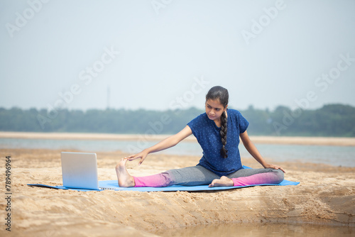 modern indian women doing Head to Knee Pose or Janu Sirsasana yoga using laptop in the sand beach