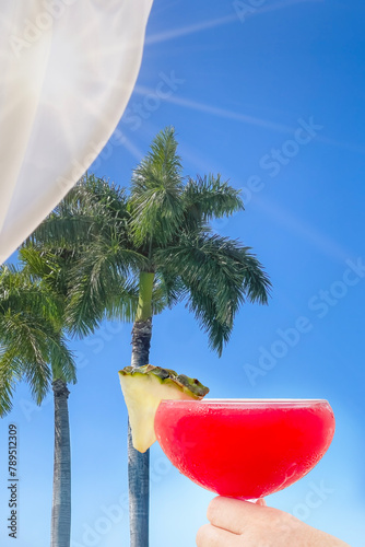 Summer sun shines through silk fabric on a cabana while a woman holds a tropical drink and looks out at palm trees photo