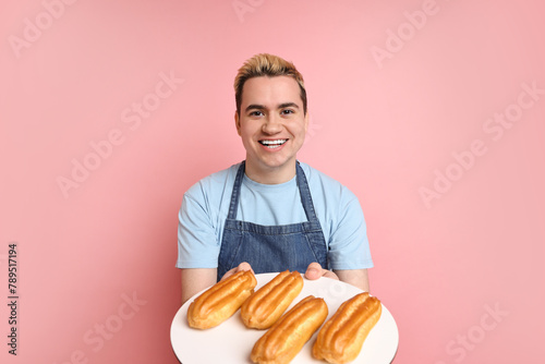 Portrait of happy confectioner holding plate with delicious eclairs on pink background