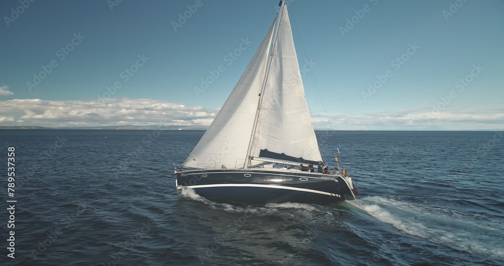 Fototapeta premium Slow motion closeup of sail luxury yacht at ocean bay aerial. Boat race at open sea. Sailboat regatta at Brodick harbor, Arran Island, Scotland, Europe. Seascape with sun reflection at summer cruise