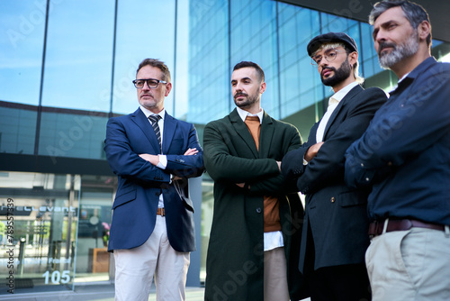 Low angle view group business men diverse ages in formal suit standing side by side with arms crossed in front of work office building posing serious for photo. Only male entrepreneur elegant people photo