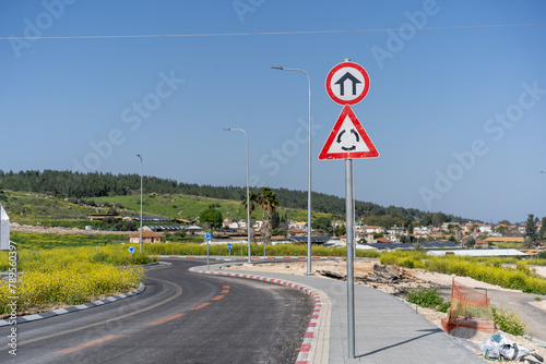 Pair of road signs against a blue sky, with the first indicating an upcoming roundabout and the second marking the start of an urban area.