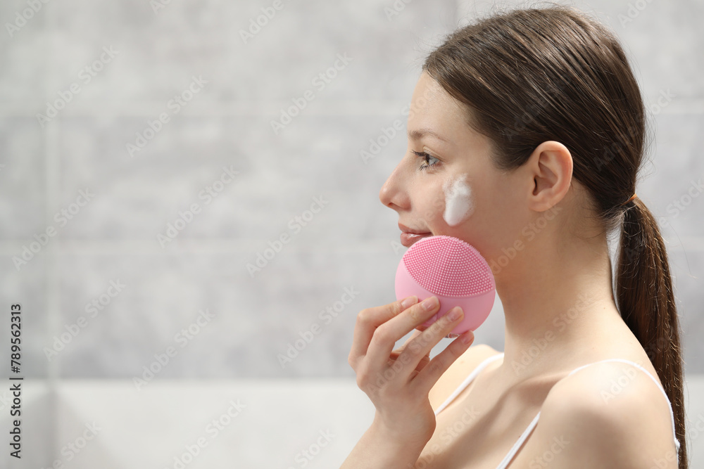 Young woman washing face with brush and cleansing foam indoors. Space for text Young woman washing face with brush and cleansing foam indoors. Space for text
