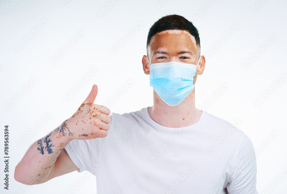 Man, portrait and thumbs up with facemask for protection from flu, illness or virus on a white studio background. Young male person or vitiligo with like emoji, yes sign or ok for health and safety Man, portrait and thumbs up with facemask for protection from flu, illness or virus on a white studio background. Young male person or vitiligo with like emoji, yes sign or ok for health and safety