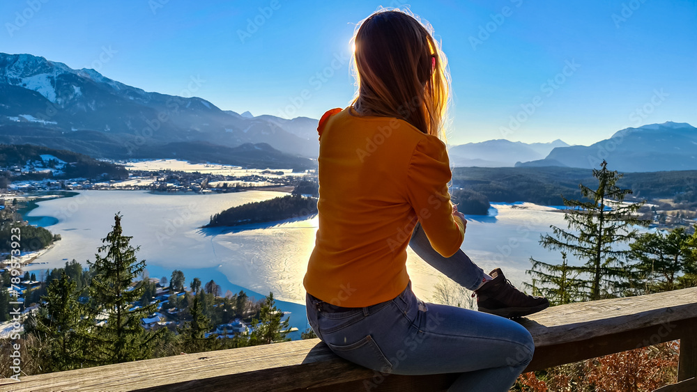 Woman sitting on railing of Taborhoehe viewing platform in Carinthia, Austria, Europe. Surrounded by high snow capped Austrian Alps mountains. lake surface is frozen. Alpine Landscape in frosty winter
