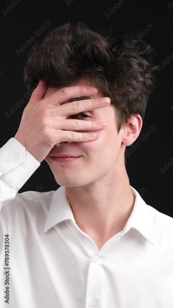Portrait of a Caucasian happy young man playfully covering eyes on black background. Romantic flirt, positive emotion concept. Vertical video. Portrait of a Caucasian happy young man playfully covering eyes on black background. Romantic flirt, positive emotion concept. Vertical video.