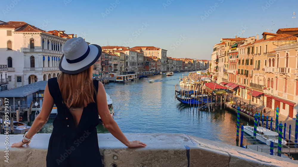 Tourist woman standing on top of famous Rialto bridge overlooking the Canal Grande in Venice, Veneto, Northern Italy, Europe. Female model is wearing black dress. Romantic luxury summer vacation