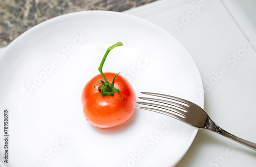 Red ripe single tomato on white porcelain plate with one metal silver fork.
