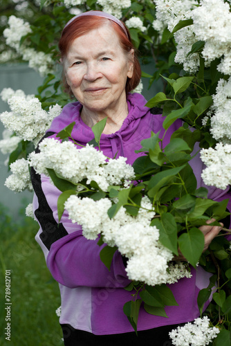 Outdoor close-up portrait of old senior woman. Beautiful elderly woman smiling against background of blooming white lilacs in spring park.