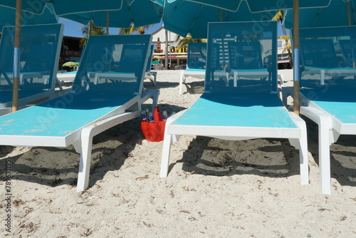 Pale blue chairs and umbrellas on the sandy beach next to which is a red plastic bucket full of empty beer bottles on Philipsburg in Sint Maarten in Caribbean Sea.