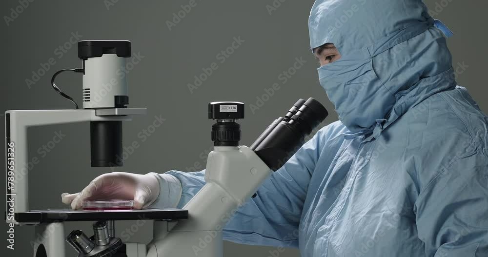 Female scientist in hazmat puts Petri dish with a sample under microscope. Analyzing sample in a Petri dish. Vaccines and medicines development Female scientist in hazmat puts Petri dish with a sample under microscope. Analyzing sample in a Petri dish. Vaccines and medicines development