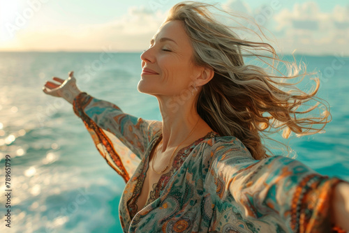 A free middle-aged woman with outstretched arms and closed eyes, dressed in an ethnic dress, enjoys the fresh air on the seashore, her hair fluttering in the wind. The concept of unity with nature