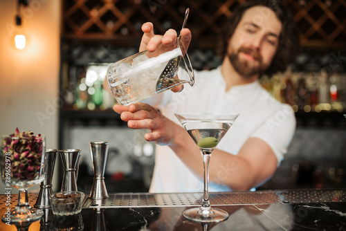 A skilled male bartender confidently pours a martini into a glass at a well-equipped, modern bar, showcasing his expertise and the sophisticated ambience.