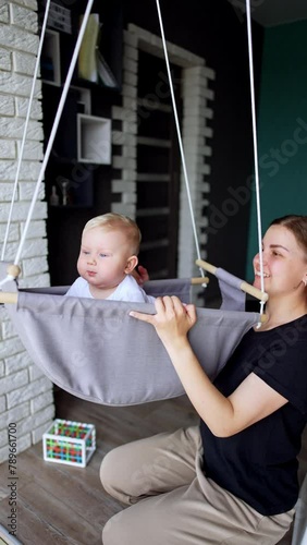 Woman holds a swing with her child sitting inside. Mother and son at home having fun time. Vertical video.