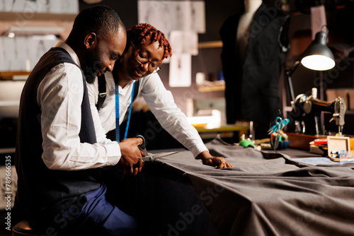 Experienced african american atelier shop coworkers meticulously cutting refined textile material for customer comissioned suit. Expert suitmakers manufacturing bespoke sartorial outfit photo