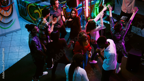 Multiethnic group of friends partying, showing funky dance moves at nightclub. Men and women dancing on electronic beats having fun on night out, enjoying colorful spotlights. Handheld shot.