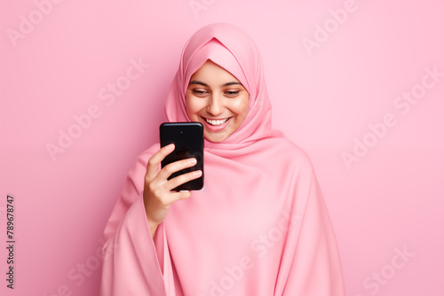 smiling arab young woman with pink veil looking at her smart phone on pink background