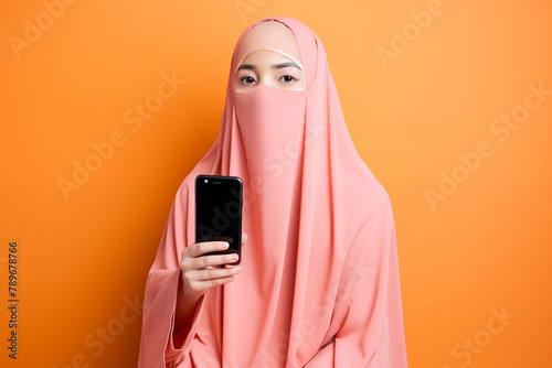 young arab woman with pink veil looking at her smart phone on orange background