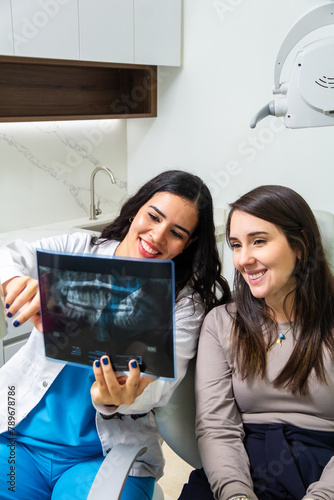 Latin female smiling dentist explaining teeth x-ray scan to caucasian woman patient sitting in a dental chair. Health care concept.