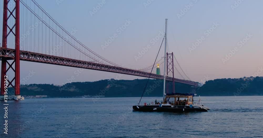 Boat ship yacht sails under big suspension metal bridge connecting the city of Lisbon and Almada, Portugal. Beautiful sunset sky panorama. Massive industrial construction. Chill water sport activities Boat ship yacht sails under big suspension metal bridge connecting the city of Lisbon and Almada, Portugal. Beautiful sunset sky panorama. Massive industrial construction. Chill water sport activities