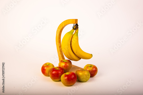 Bananas hanging from stand with apples around base on white seamless background.