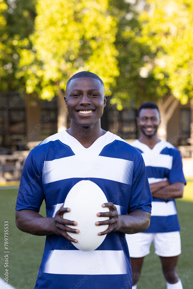 Fototapeta premium Two African American young male athletes are holding a rugby ball, smiling, on a field outdoors