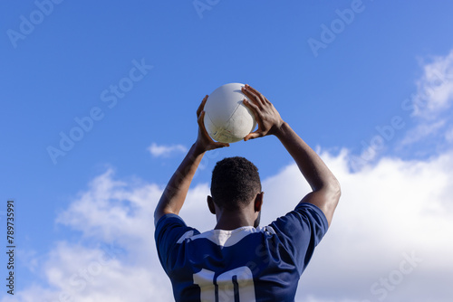 African American young male athlete holding white rugby ball above head on field, copy space
