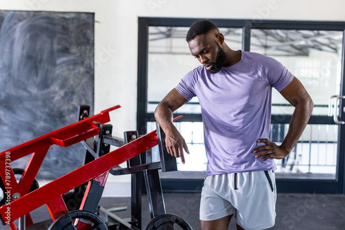 African American young male athlete standing next to gym equipment in gym, looking thoughtful