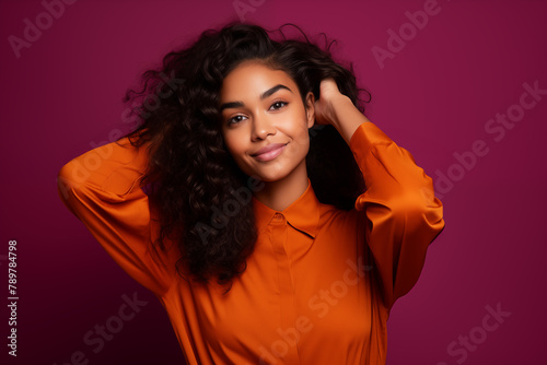 A woman with voluminous curls smiles radiantly  hands playfully in her hair  wearing a vibrant orange top against a rich magenta background.