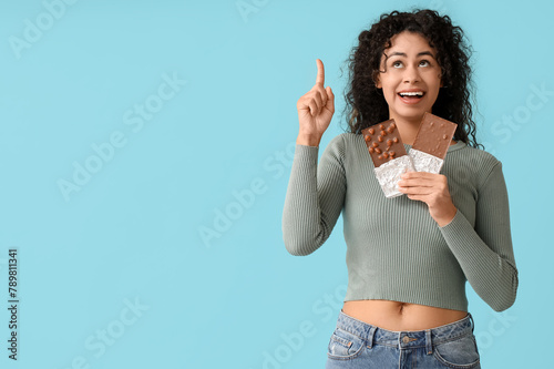 Beautiful young African-American woman with sweet chocolate bars pointing at something on blue background © Pixel-Shot