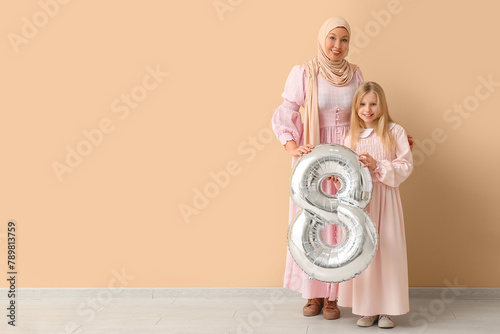 Beautiful young Muslim woman and her cute little daughter with air balloon in shape of figure 8 near beige wall