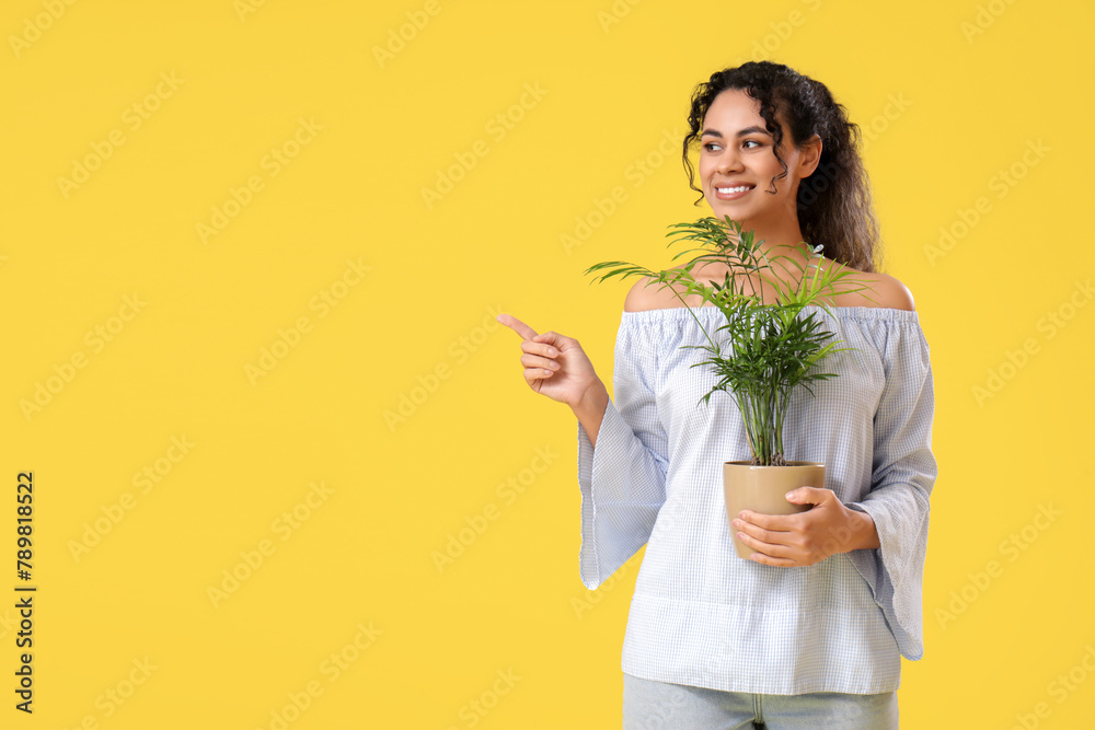 Smiling beautiful young African-American woman with plant in her hand pointing at something on yellow background Smiling beautiful young African-American woman with plant in her hand pointing at something on yellow background
