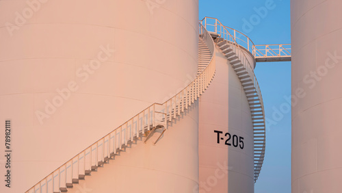 Curve lines pattern of spiral staircase on group of white storage fuel tanks with orange sunlight on surface against blue evening sky background