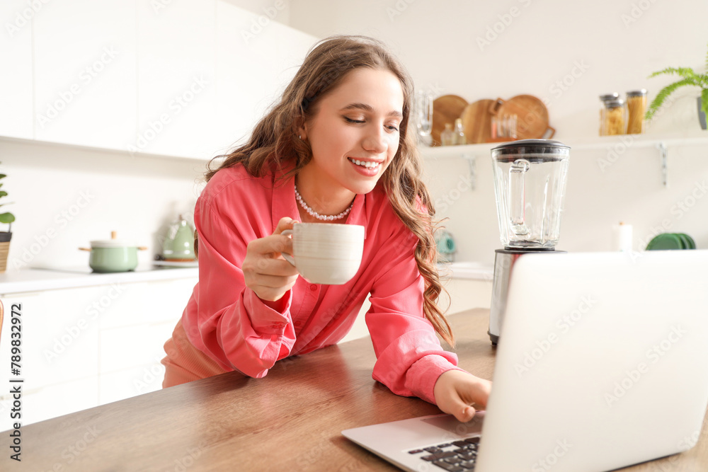 Beautiful young woman with coffee cup using laptop at table in kitchen Beautiful young woman with coffee cup using laptop at table in kitchen