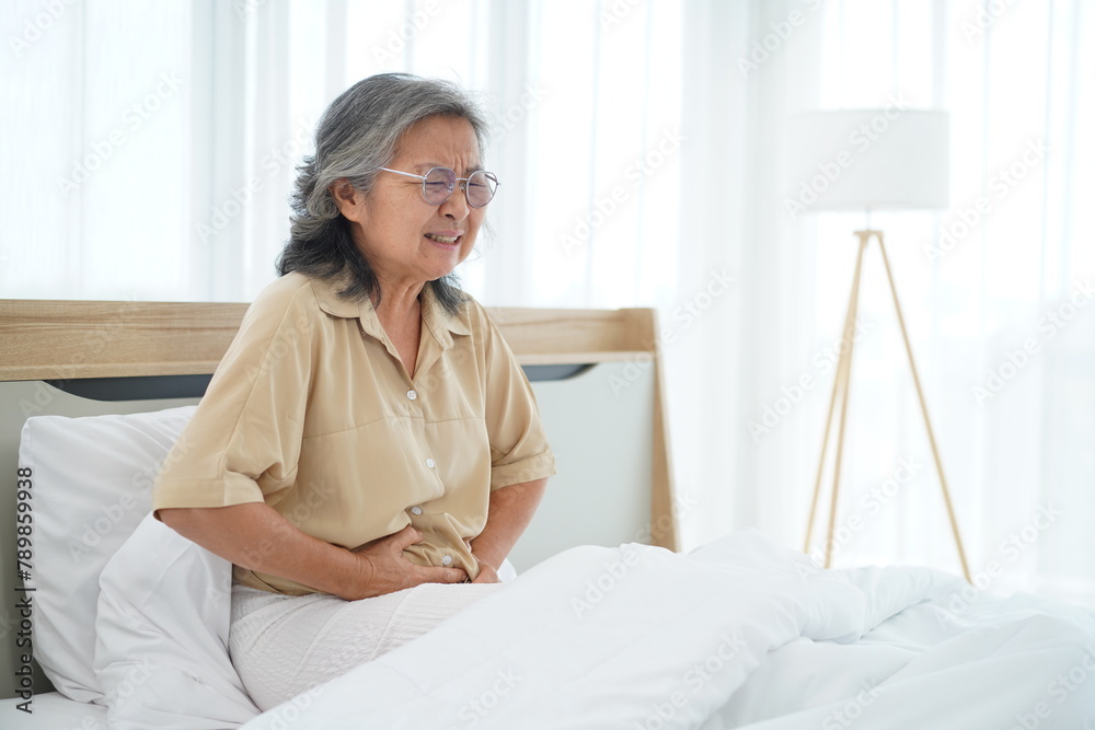Mature Woman sitting on bed and holding her stomach with feels stomachache Mature Woman sitting on bed and holding her stomach with feels stomachache