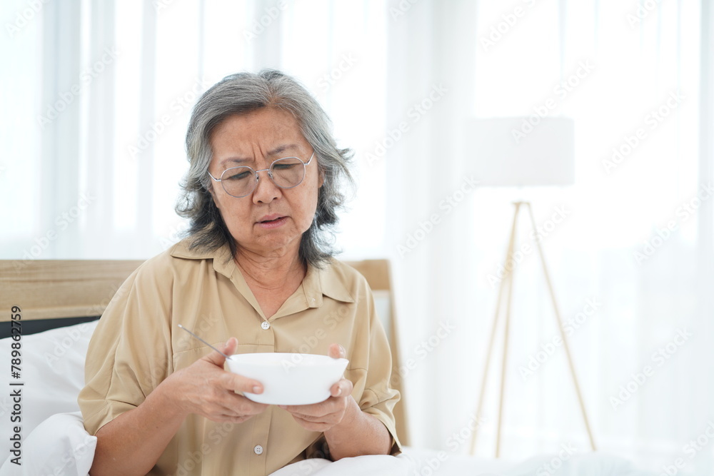 Senior woman wearing glasses sitting on bed hold a bowl with feeling anorexic Senior woman wearing glasses sitting on bed hold a bowl with feeling anorexic