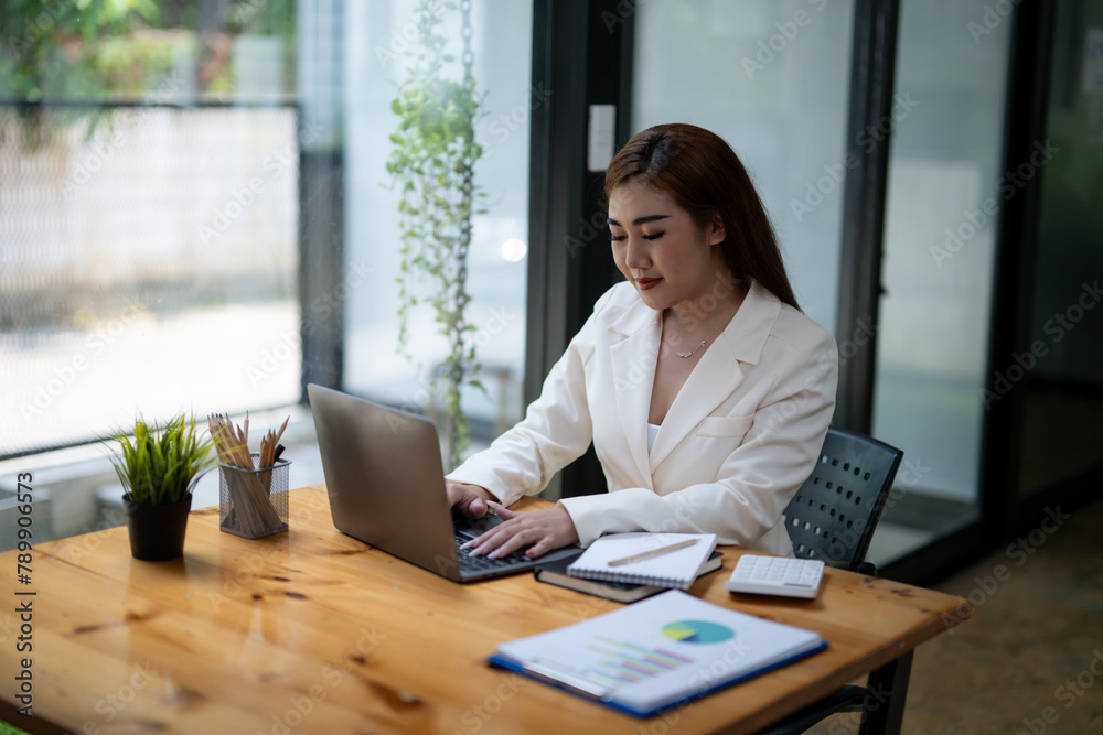 Professional Asian businesswoman typing on laptop at a wooden desk with paperwork and greenery in a bright office setting. Professional Asian businesswoman typing on laptop at a wooden desk with paperwork and greenery in a bright office setting.
