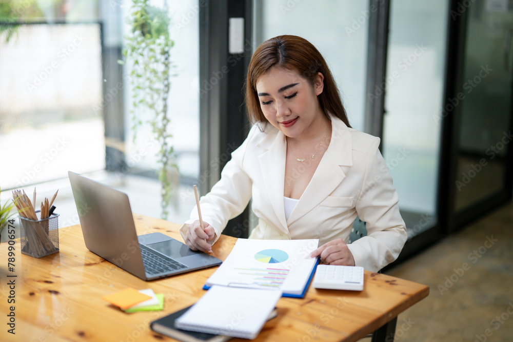 Professional Asian businesswoman typing on laptop at a wooden desk with paperwork and greenery in a bright office setting. Professional Asian businesswoman typing on laptop at a wooden desk with paperwork and greenery in a bright office setting.