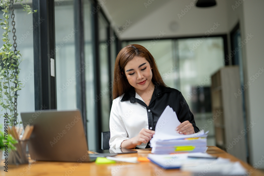 Focused businesswoman in a two-tone shirt sorting through stacks of paperwork at a wooden desk in an office environment. Focused businesswoman in a two-tone shirt sorting through stacks of paperwork at a wooden desk in an office environment.