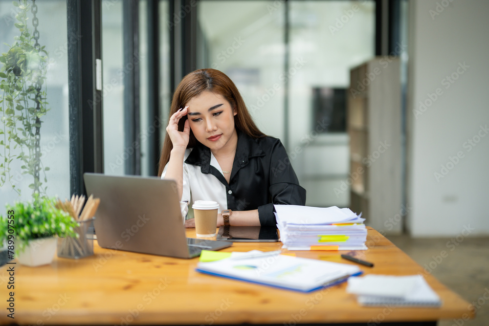 Pensive Asian businesswoman reading a report, with a laptop and paperwork on her desk in a bright office space. Pensive Asian businesswoman reading a report, with a laptop and paperwork on her desk in a bright office space.