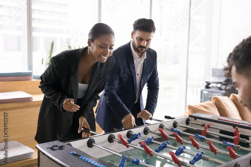 Positive excited young multiethnic couple of professionals playing toy football, competing in table soccer with coworkers, turning handles at field miniature, laughing, relaxing on work break © fizkes