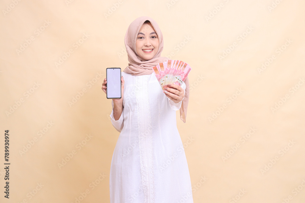 woman indonesia young wearing a hijab smiles cheerfully holding a cell phone gadget and rupiah money in her hand. Lifestyle, business and commercial concept woman indonesia young wearing a hijab smiles cheerfully holding a cell phone gadget and rupiah money in her hand. Lifestyle, business and commercial concept