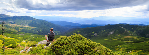 Man and dog thoughtfully enjoying the mountain landscape view of the valley. Beautiful green landscape. Friendship and loneliness concept. Long horizontal banner wild nature