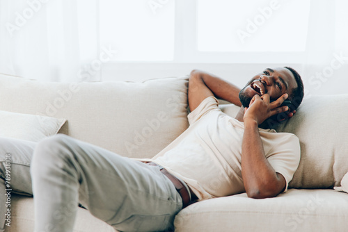 Happy African American Man Talking on Black Smartphone while Sitting on a Modern White Sofa at Home He is Engaged in a Video Call, Typing Messages and Using Social Media The Man appears Relaxed and