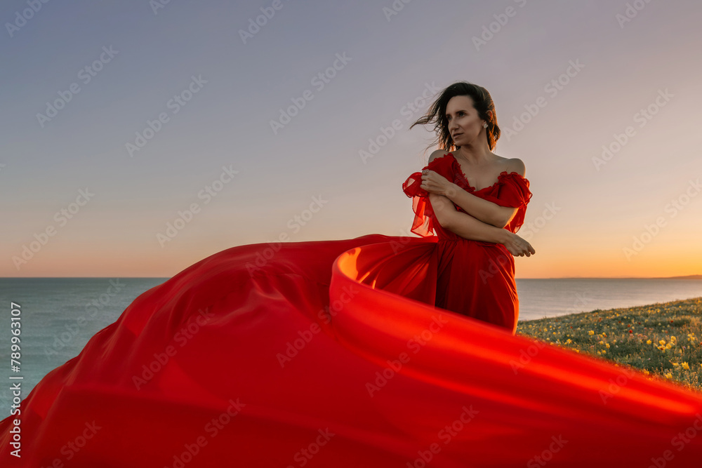 A woman in a red dress is standing on a grassy hillside. The sun is setting in the background, casting a warm glow over the scene. The woman is enjoying the beautiful view and the peaceful atmosphere.