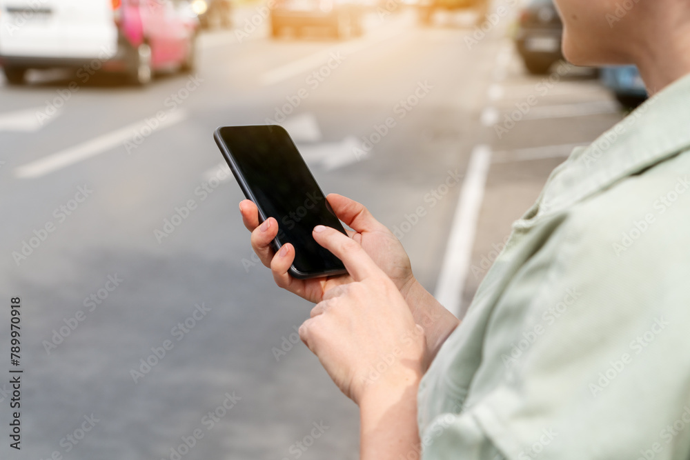 Modern female person stands next to road in city and using her smartphone. Searching car rental, taxi, public transportation.