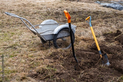 A wheelbarrow and shovel rest in the dirt of a grassy field