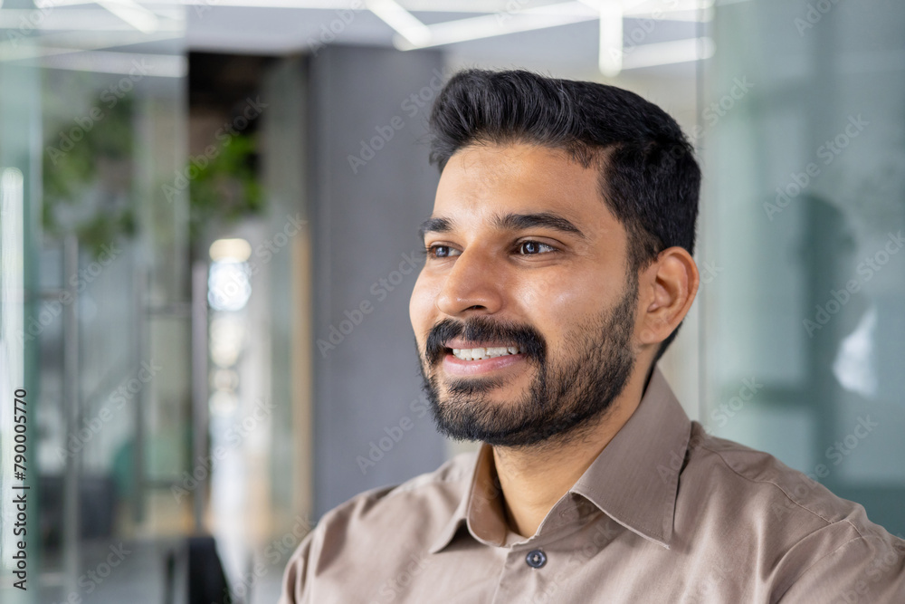 Obraz premium A portrait of a confident young man with a beard smiling warmly in a well-lit, contemporary office environment. The image conveys a sense of happiness and professional success.