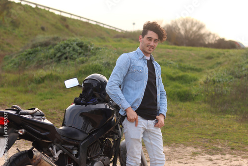 Young Caucasian man with curly hair standing next to the motorcycle with hands in pockets looking at camera smiling