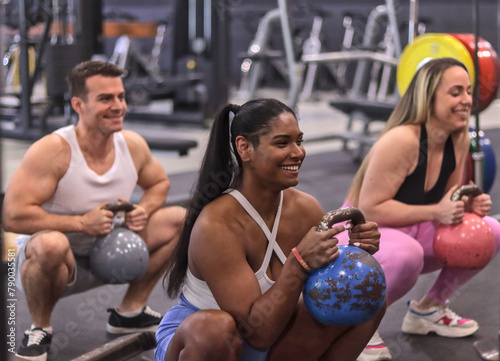 Group of multiracial smiling people doing squats with kettle bell dumbbells, in a gym, sport dress. Fitness and healthy lifestyle concept